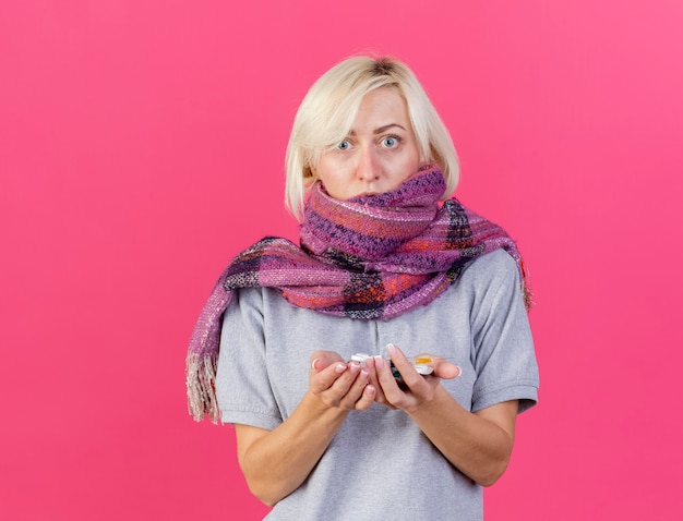 Die besorgte junge blonde kranke slawische Frau, die Schal trägt, hält Packungen von medizinischen Pillen