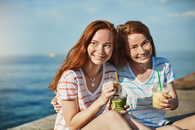 Die besten Momente teilen Zwei wunderschöne Mädchen mit roten Haaren und Sommersprossen sitzen in der Nähe der Küste und trinken Cocktails, umarmen sich und genießen es, Zeit miteinander zu verbringen, sich entspannt und glücklich zu fühlen