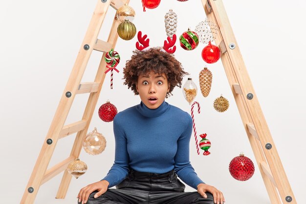 Die betäubte dunkelhäutige Frau sitzt mit gekreuzten Beinen über der Leiter mit Weihnachtsspielzeug und bereitet sich auf die Winterferienfeier vor. Weihnachtsdekoration. Was für eine große Überraschung