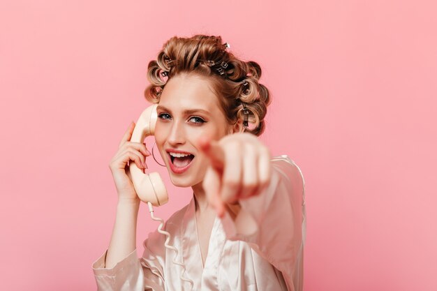 Die blonde Frau im Seiden-Home-Outfit telefoniert und zeigt mit dem Finger nach vorne gegen die isolierte Wand