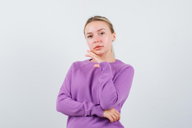 Die blonde Frau schaut in die Kamera, indem sie ihre Hand am Kinn auf weißem Hintergrund hält