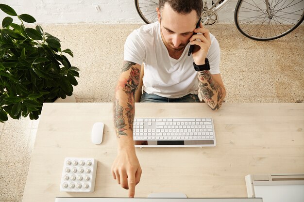 Die Draufsicht des tätowierten Mannes zeigt etwas auf dem Display, während er auf seinem Arbeitsdesktop im Co-Working-Center telefoniert