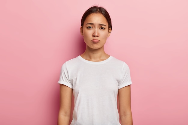 Die düstere, traurige Asiatin sieht unglücklich aus, drückt Bedauern und Traurigkeit aus, runzelt die Stirn, ist frustriert, trägt ein weißes T-Shirt und zeigt negative Emotionen, die durch schlechte Nachrichten über unfaires Leben verärgert sind