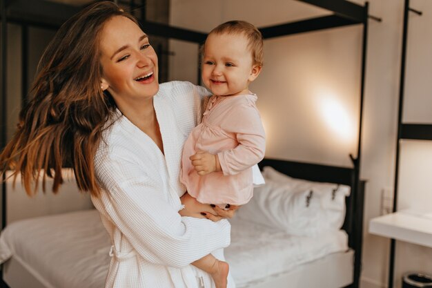 Die dunkelhaarige Dame im weißen Bademantel und ihre kleine Tochter lachen aufrichtig, während sie im hellen Schlafzimmer spielen.