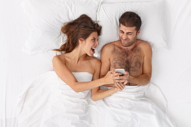 Die emotionale Frau ruft aus und zeigt dem Ehemann etwas auf dem Handy, bleibt zusammen im Bett, ruht sich morgens aus. Junge Ehepartner, die von modernen Technologien abhängig sind, lesen Nachrichten oder sehen sich online Videos an.