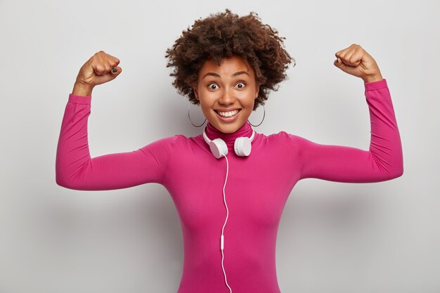 Die energiegeladene afroamerikanische Frau hebt die Arme, um Muskeln und Kraft zu zeigen, lächelt breit und trägt Stereokopfhörer um den Hals