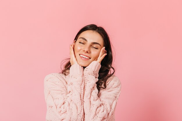 Die entzückende brünette Frau lächelt zärtlich mit geschlossenen Augen. Dame im warmen Pullover, der im rosa Studio aufwirft.