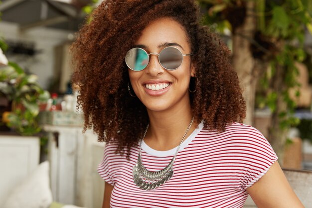 Die entzückende, dunkelhäutige, lächelnde, süße Frau mit Afro-Frisur trägt eine modische Sonnenbrille, genießt die Sommererholung, sitzt gegen das Café-Interieur und hat ein angenehmes Gespräch mit einem Freund, der gegenüber sitzt