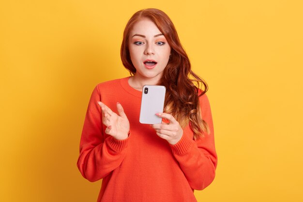 Die fassungslose europäische Frau steht unter Schock, liest schreckliche Nachrichten aus dem Mobilfunkgerät, hat welliges rotes Haar und trägt einen orangefarbenen Pullover