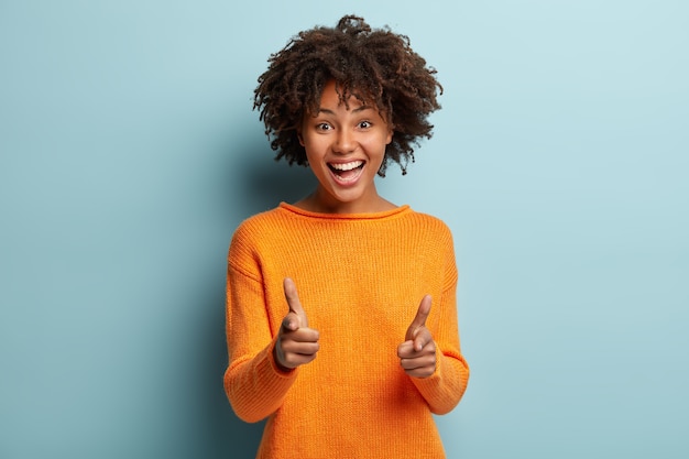 Die freudige hübsche Afroamerikanerin macht eine Fingerpistolengeste in die Kamera, drückt ihre Wahl aus, lächelt breit und trägt einen orangefarbenen Pullover