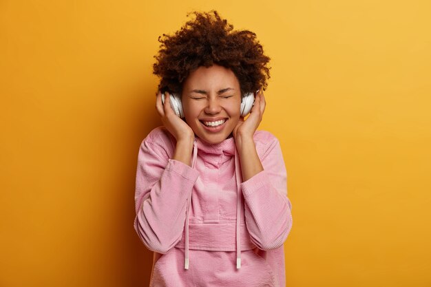 Die fröhliche Frau mit den lockigen Haaren genießt jedes Lieblingslied, hört Musik in Stereokopfhörern, schließt die Augen und lächelt breit, inspiriert von großartiger Musik, gekleidet in Freizeitkleidung, steht drinnen