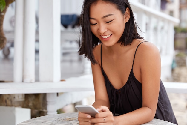 Die fröhliche Japanerin installiert eine neue Anwendung auf dem Handy, während sie auf ein Gericht im Restaurant wartet, das mit dem drahtlosen Internet verbunden ist. Menschen-, Freizeit- und Technologiekonzept