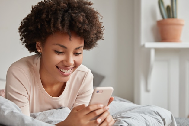 Die fröhliche schwarze afroamerikanische Frau liegt mit einem modernen Handy im Bett, liest angenehme Kommentare unter ihrem Foto in sozialen Netzwerken über ein Smartphone, genießt die Schlafenszeit und das Internet allein in einem gemütlichen Raum