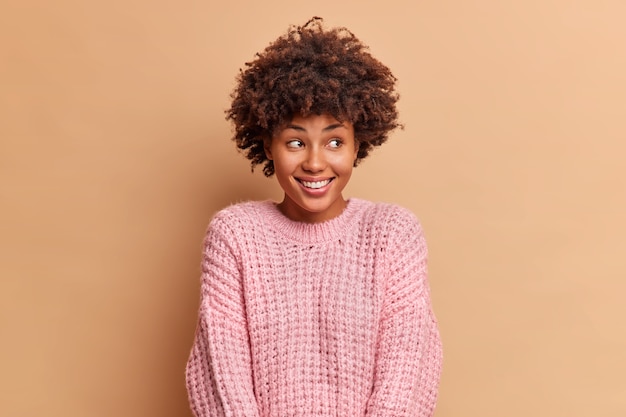 Die fröhliche tausendjährige frau mit afro-haaren lächelt sanft und schaut weg, wenn sie in hochstimmung ist. sie drückt positive emotionen aus, die über der beige studiowand isoliert sind