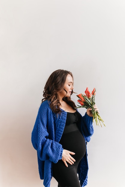 Die gelockte glückliche schwangere Frau in der blauen Strickjacke hält Blumen. Charmantes brünettes Mädchen im schwarzen Kleid riecht Tulpenstrauß auf weißem Hintergrund.