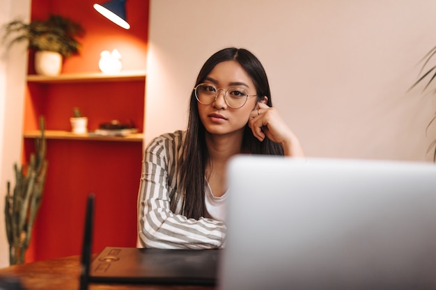 Die Geschäftsdame in der gestreiften Jacke und in der Brille schaut nach vorne und lehnt sich an den Tisch gegen den Laptop