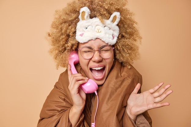 Die gesellige junge frau mit lockigem haar ruft laut aus, benutzt ein retro-telefon, hält den hörer in der nähe des ohrs, trägt eine schlafmaske auf der mit einer decke bedeckten stirn und hält die erhobenen handflächen laut isoliert an der braunen wand