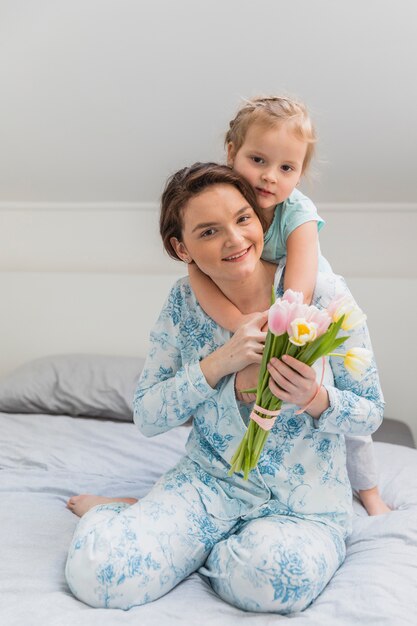 Die glückliche junge Frau, die piggyback ihrer kleinen Tochter Fahrt auf das Bett hält, das Bündel Tulpenblumen gibt, blüht