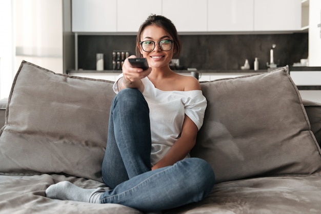 Die glückliche junge Frau, die zu Hause auf Sofa sitzt, sehen fern.