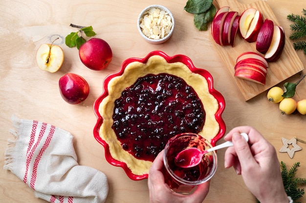 Die Hände der Frauen halten ein Glas Marmelade Der Prozess der Herstellung eines Apfelkuchens oder -kuchens auf einem Holztisch Apfelkuchen mit Preiselbeermarmelade und Backzutaten Apfelkuchen für Neujahr und Weihnachten