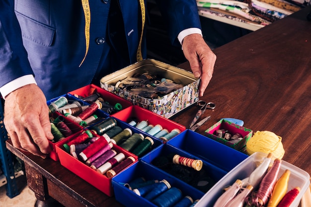 Die Hände des männlichen Modedesigners auf dem Behälter, der unterschiedliche Art von Threadspulen auf Holztisch enthält