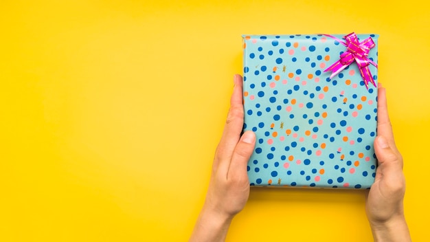 Die Hände, die Tupfen halten, wickelten Geschenkbox mit rosa Bogen auf gelbem Hintergrund ein