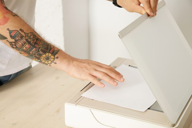 Die Hände öffnen ein Scannerfach und legen ein Blatt Papier ein, um ein Dokument in einem multifunktionalen elektronischen Gerät zu scannen, das auf einem weißen Holztisch isoliert ist.