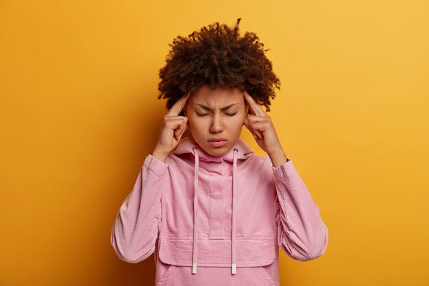 Die halbe Einstellung einer verzweifelten dunkelhäutigen Frau verspürt schmerzhafte Migräne oder Kopfschmerzen, berührt Schläfen und blinzelt das Gesicht, ist nach einem müden Treffen müde, versucht sich zu konzentrieren, trägt ein rosa Sweatshirt