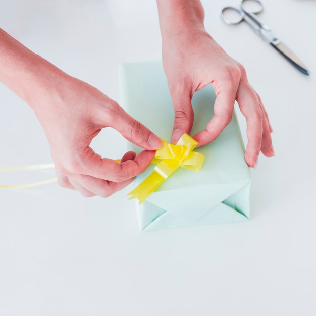 Kostenloses Foto die hand der frau, die das gelbe band auf eingewickelter geschenkbox über weißem schreibtisch haftet