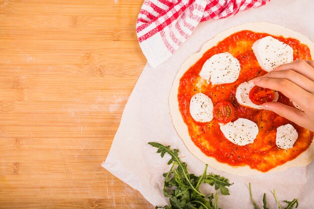 Die Hand der Frau, die Käse und Tomate auf Teig für das Vorbereiten der Pizza setzt