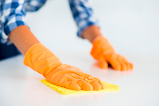 Die Hand der Frau, die orange Handschuhe trägt, wischt den weißen Schreibtisch mit gelbem Staubtuch ab