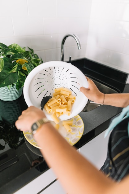 Die hand der frau, die rigatoni-teigwaren vom weißen sieb auf platte setzt