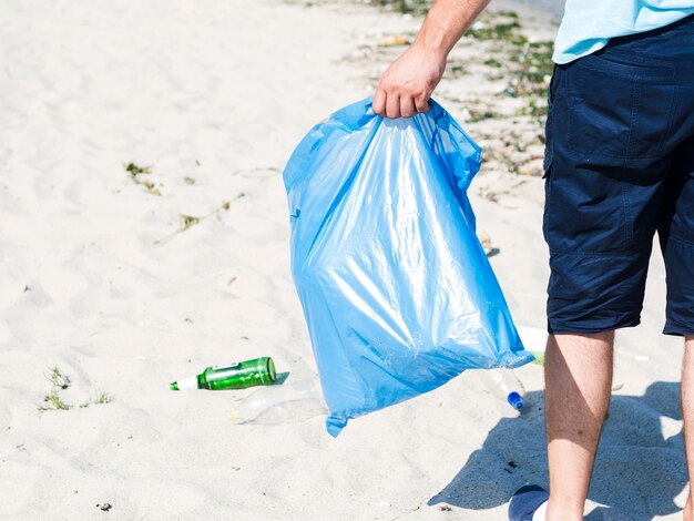 Die Hand des Mannes, die blaue Abfalltasche auf Strand trägt