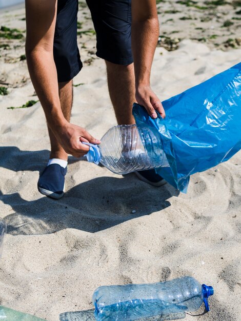 Die Hand des Mannes, die Plastikflasche in blauen Abfallbeutel auf Strand einsetzt