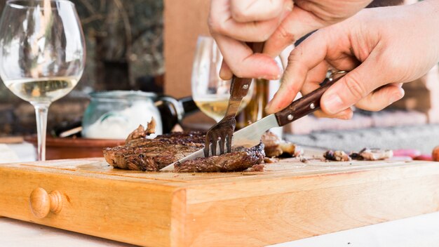 Die Hand des Mannes, die Rindfleischsteak mit Gabel und Messer auf hackendem Brett mit Fach schneidet