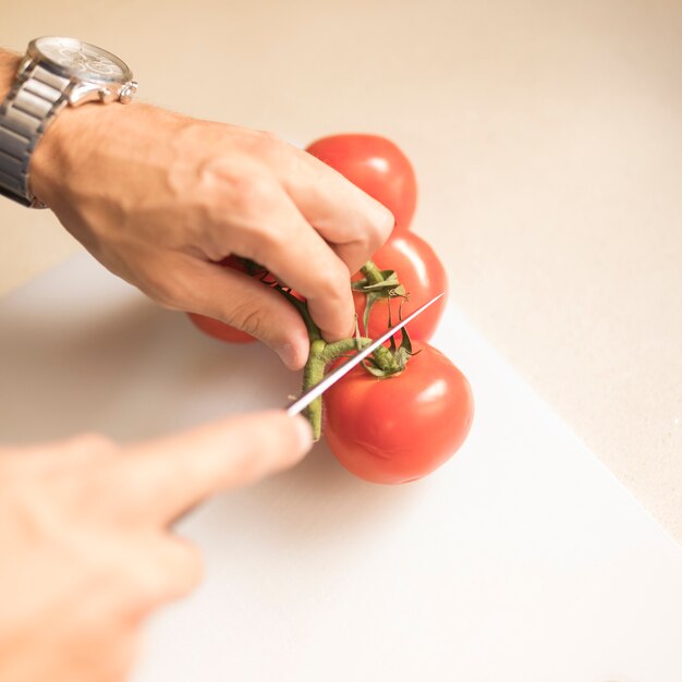 Die Hand des Mannes, die Stamm der roten Tomate mit scharfem Messer auf hackendem Brett schneidet