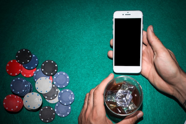 Kostenloses Foto die hand des pokerspielers, die handy zeigt und whiskyglas auf schürhakentabelle hält