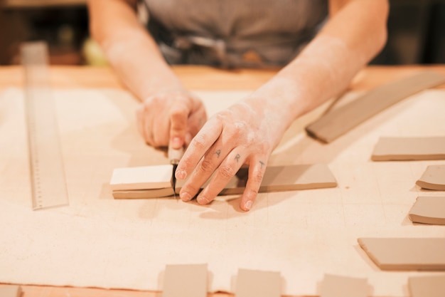 Die Hand des weiblichen Töpfers, die den Lehm in der Fliesenform mit Werkzeug auf hölzernem Schreibtisch schneidet