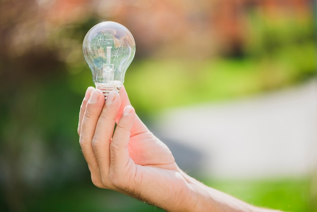 Die Hand eines Mannes, die transparente Glühlampe gegen unscharfen Hintergrund zeigt