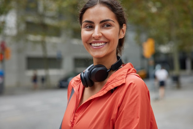 Die horizontale Aufnahme einer brünetten Frau mit fröhlichem Gesichtsausdruck führt zu einem aktiven Lebensstil, der in einer Windjacke gekleidet ist und Stereo-Kopfhörer trägt, die draußen vor verschwommenem Hintergrund stehen Sportkonzept