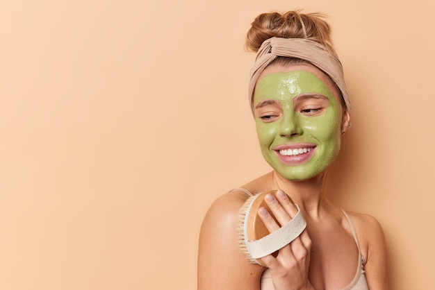 Die horizontale Aufnahme einer erfreuten jungen Frau wendet eine grüne Pflegemaske auf dem Gesicht an, verwendet eine Körperbürste, steht mit nackten Schultern isoliert auf braunem Hintergrund mit Kopierfläche für Ihre Werbeinhalte