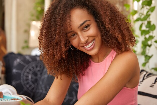 Die horizontale Aufnahme einer fröhlichen dunkelhäutigen Frau hat knackiges Haar, trägt eine lässige rosa Weste, lächelt breit, posiert auf der Terrasse auf der Couch, drückt positive Emotionen aus und hat Freizeit