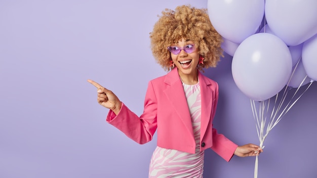 Die horizontale Aufnahme einer fröhlichen Frau in modischer Kleidung hält einen Haufen aufgeblasener Luftballons, feiert den Universitätsabgang und organisiert Partypunkte auf einem Kopierraum, der auf violettem Hintergrund isoliert ist