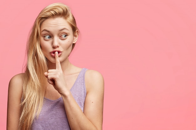 Die horizontale Aufnahme einer geheimen blonden jungen Frau zeigt ein stilles Zeichen, hält den Vorderfinger auf den Lippen und schaut auf mysteriöse Weise auf eine leere Kopie