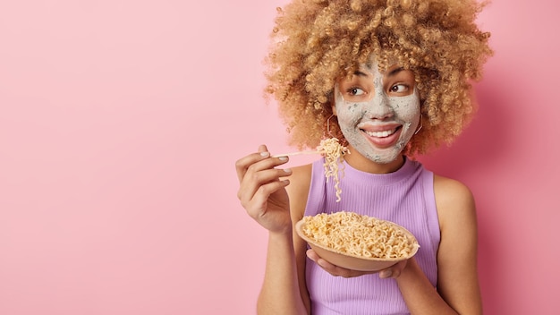 Die horizontale Aufnahme einer gut aussehenden jungen Frau mit lockigem Haar trägt eine Schönheitsmaske auf dem Gesicht auf, die sich Hautpflegebehandlungen unterzieht, isst köstliche Spaghetti und trägt ein lässiges T-Shirt, das über rosafarbenem Hintergrund isoliert ist