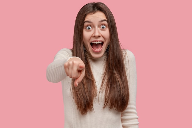 Die horizontale Aufnahme einer gut aussehenden positiven jungen Frau zeigt mit dem Zeigefinger direkt auf die Kamera, hält den Mund weit geöffnet und trägt Freizeitkleidung