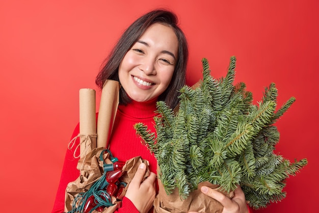 Die horizontale Aufnahme einer hübschen brünetten Asiatin mit sanftem Lächeln im Gesicht neigt den Kopf in Posen mit Urlaubsattributen und bereitet sich isoliert auf leuchtend rotem Hintergrund auf das neue Jahr vor. Weihnachten steht vor der Tür