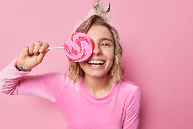 Die horizontale Aufnahme einer positiven jungen hübschen Frau mit Frisur bedeckt das Auge mit runden Karamellbonbons, trägt eine Krone und festliche Kleidung. Das Lächeln zeigt breit weiße Zähne, die über rosafarbenem Hintergrund isoliert sind