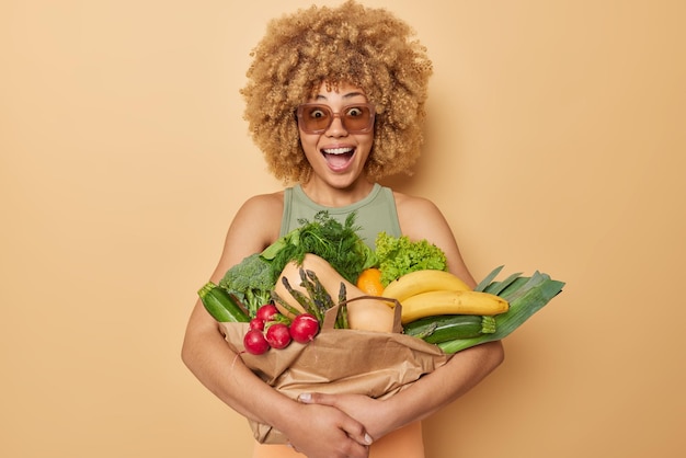 Kostenloses Foto die horizontale aufnahme einer überraschten, lockigen jungen frau umarmt frisches gemüse und obst, die in papier eingewickelt sind, und reagiert auf etwas unerwartetes, das lebensmittel einzeln auf braunem hintergrund trägt