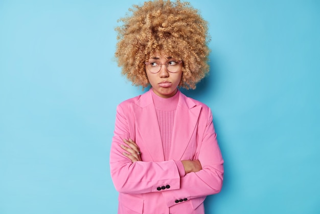 Kostenloses Foto die horizontale aufnahme einer verärgerten, düsteren frau mit lockigem haar hält die arme verschränkt, sieht beleidigt und unzufrieden aus, hört schlechte worte und trägt eine transparente brille in rosafarbenem, formellem outfit, die über blauem hintergrund isoliert ist